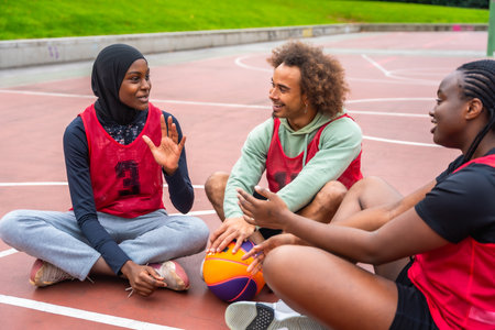 Group of young adult friends from different backgrounds sitting on a basketball court, taking a break and engaging in conversation, sharing a moment of camaraderie and connectionの写真素材