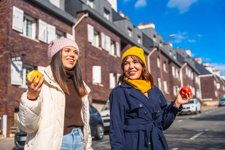 Two smiling young women friends are enjoying a sunny winter day, walking along a city street and holding fresh apples, promoting healthy eating and an active urban lifestyleの写真素材