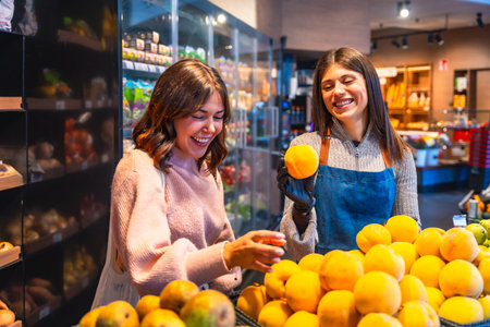 Female customer happily selects fresh oranges from a bright produce display while a cheerful apron clad clerk assists, showcasing friendly service and healthy food choicesの写真素材