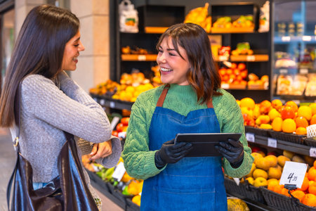 Friendly store employee wearing an apron and gloves holding a digital tablet, helping a smiling...
