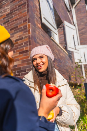 Two young women chatting outdoors on a sunny urban street, smiling and sharing apples while bundled in warm clothing, enjoying friendship, fresh food and relaxed conversationの写真素材