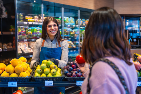 Smiling woman greengrocer assisting a customer choosing fresh organic fruit, including pears, plums, and papayas, displayed in baskets at a modern grocery storeの写真素材