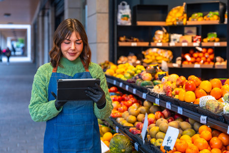 Female small business owner wearing an apron and gloves managing stock and orders on a digital tablet while standing in front of a colorful fruit and vegetable displayの写真素材