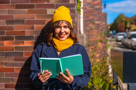 Young woman wearing a yellow beanie and scarf, dressed in a trench coat, standing outdoors in warm sunny autumn light, reading an open book against a brick wallの写真素材
