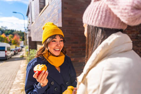 Two cheerful women friends enjoying fresh apples and engaging in a conversation outdoors on an urban street during a bright autumn day, reflecting healthy eating and friendshipの写真素材