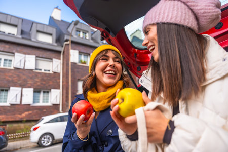 Two cheerful young women laughing while wearing hats and holding apples, standing by an open car trunk in an urban setting during cold weather, enjoying friendshipの写真素材