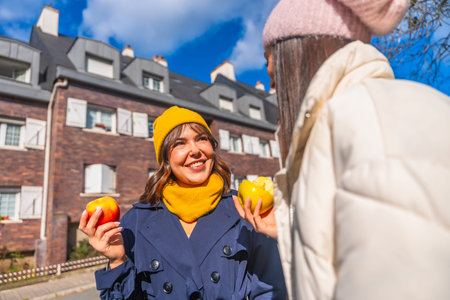 Two young women laugh and chat on a sunny autumn city street, sharing fresh apples and cozying up in...
