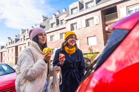 Two young adult women friends enjoying a healthy snack of apples while walking on a city street, embracing a sustainable and active urban lifestyle in a cheerful momentの写真素材