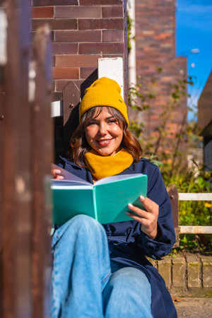 Young woman in a yellow beanie sits on a bench against a brick wall, smiling and reading a book while enjoying warm autumn sunlight in a relaxed urban settingの写真素材