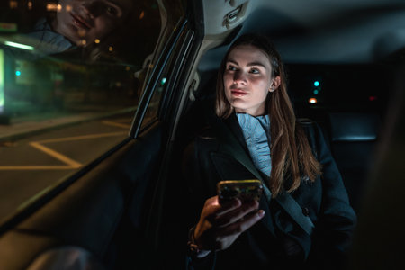 Woman traveling in the back seat of a car at night, holding a smartphone and looking out the window with city lights reflecting on the glass, representing urban transportation servicesの写真素材