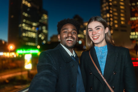 Diverse young man and woman smiling happily while taking a selfie together outdoors at night, with a modern glowing city skyline creating a soft bokeh backgroundの写真素材