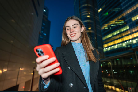 Young woman smiling outdoors at night, interacting with a smartphone in an urban environment with office buildings illuminated in the background, representing communication and social networkingの写真素材