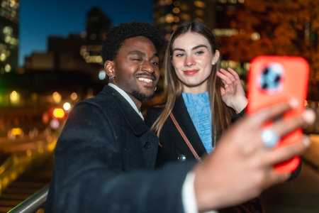 Diverse young couple on a city street at night, smiling and taking a candid selfie with a smartphone during a joyful, modern date night outdoors, sharing a happy momentの写真素材
