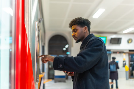 Young black man standing at a ticket machine in a modern train station, purchasing a ticket for his daily commute or a business trip, reflecting urban transportation and travel conceptsの写真素材