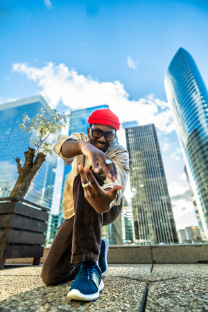 Young black man with a red beanie and glasses smiling at the camera, posing and expressing himself with dynamic movements in an urban setting surrounded by modern skyscrapersの写真素材