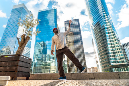 Happy black man celebrating joy and freedom with a dynamic dance on an urban plaza, surrounded by towering contemporary skyscrapers under a blue sky, symbolizing success and aspirationの写真素材