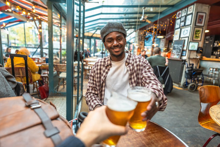 Young man smiling and clinking beer glasses with a friend in a casual pub, celebrating friendship, laughter and relaxed weekend vibes in an urban hangoutの写真素材