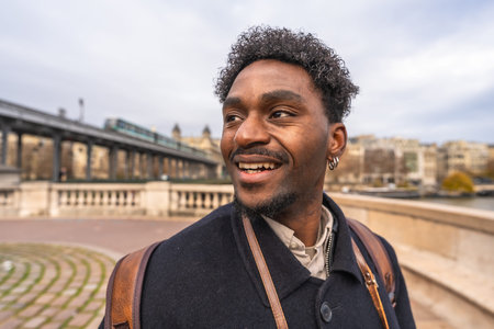 Happy black man smiling and looking away while strolling across paris bir hakeim bridge with a metro...