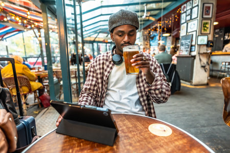 Young man in a plaid shirt and hat relaxing at a pub table, drinking a beer while browsing on a digital tablet with headphones around his neck, luggage beside himの写真素材
