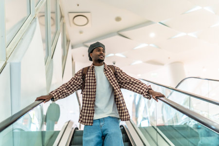 Young black man riding a modern escalator in a downtown shopping mall, wearing a backpack and stylish casual clothes, looking ahead while exploring urban retail spacesの写真素材