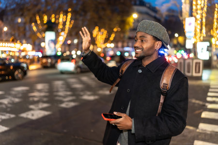 Young man smiling while hailing a taxi on a vibrant city street at night, holding a smartphone and wearing a backpack and hat, surrounded by blurred evening lightsの写真素材
