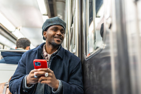 Young black man in beret and jacket rides the subway holding a red smartphone, smiling and looking...