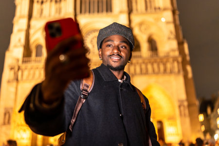Young black man with a backpack smiling and taking a selfie with his smartphone in front of the illuminated Notre Dame cathedral in Paris, enjoying a night out while travelingの写真素材