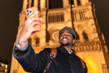 Young man smiling and taking a selfie with a smartphone, capturing a memorable moment during a night cityscape tour of the iconic Notre Dame cathedral in Paris, Franceの写真素材