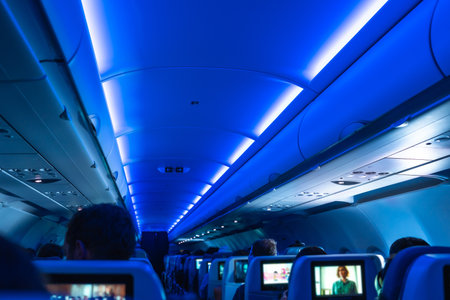 Airplane cabin illuminated with blue ambient light, passengers sitting on their seats, some watching movies on the screens embedded in the headrestsの写真素材