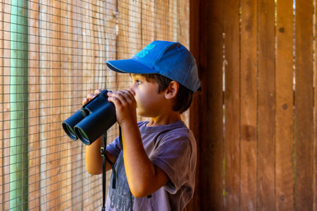 Young boy wearing a blue cap observing wildlife through binoculars at a nature park in Kamloops,...