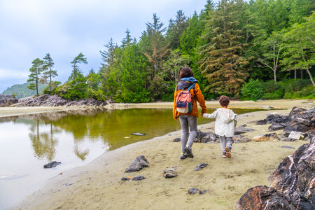 Mother and child enjoying a leisurely stroll along a sandy beach in Tofino, British Columbia, surrounded by lush forests and calm reflective watersの写真素材