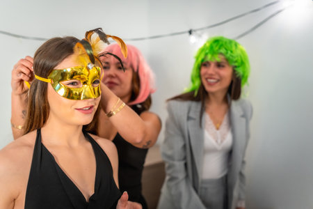 Friends preparing for a celebration, with one woman helping another put on a gold masquerade mask with feathers while others wear colorful wigs, enjoying the festive atmosphereの写真素材