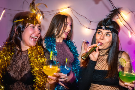 Three diverse young women in festive costumes laughing and toasting with colorful cocktails and blowing party horns beneath string lights at an indoor new years eve or costume partyの写真素材