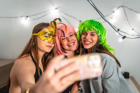 Group of happy young women celebrating new year's eve or a costume party indoors, smiling and posing for a cheerful selfie while wearing colorful wigs and a masquerade maskの写真素材
