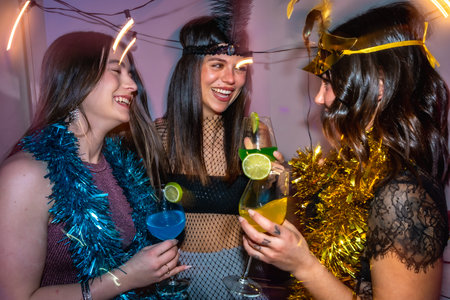 Young women friends laughing and cheering with colorful cocktails, celebrating together during a festive new year's eve or costume party with tinsel decorationsの写真素材
