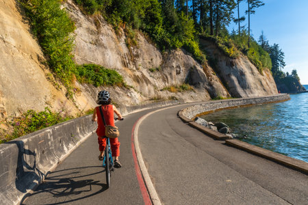 Tourist wearing helmet and carrying bag is riding bicycle along Stanley Park seawall in Vancouver, British Columbia, with cliffs and ocean in background on sunny summer dayの写真素材