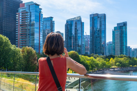 Tourist is making a phone call while enjoying the view of the modern buildings of Vancouver's skyline from the bridge on a beautiful sunny dayの写真素材