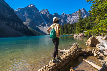 Female tourist standing on a log and admiring the turquoise waters of Moraine Lake, framed by the stunning peaks of the Canadian Rockies on a sunny summer dayの写真素材