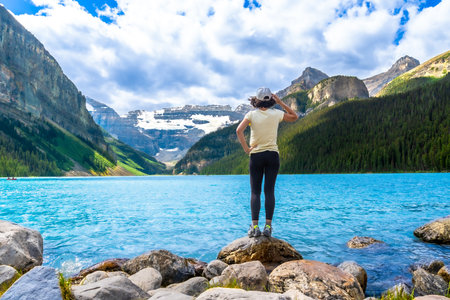 Tourist standing on rocks by the turquoise water of Lake Louise admires the surrounding Canadian rockies in Banff National Park, Alberta, on a sunny day with some cloudsの写真素材