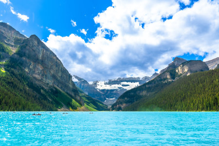 Canoes are paddling on a beautiful turquoise lake with surrounding mountains and glaciers in Banff National Park, Alberta, Canada, on a sunny summer day with white clouds and blue skyの写真素材