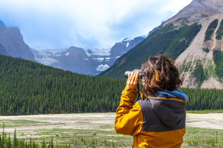 Female hiker wearing a yellow jacket using binoculars while observing a glacier in the Canadian Rockies along the Icefields Parkway, surrounded by lush pine forest and mountainsの写真素材