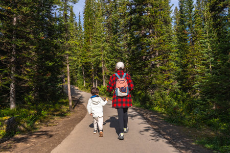 Mother and child holding hands, enjoying a leisurely hike along a paved path, surrounded by the lush green trees of Banff National Park in the Canadian Rockiesの写真素材
