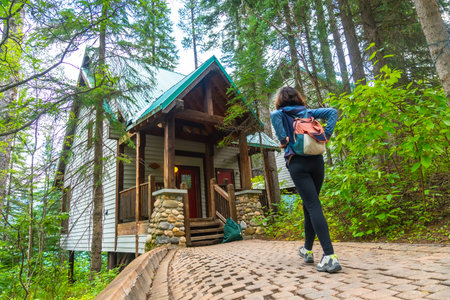 Female tourist with backpack walking up cobblestone path to a wooden cabin surrounded by lush green...