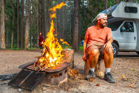 Camper enjoying warmth of campfire in Banff National Park, Canadian Rockies, with campervan parked nearby and another camper walking in the backgroundの写真素材