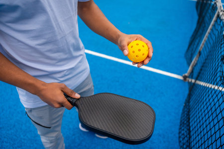Person standing on a bright blue pickleball court, holding a black paddle and a yellow perforated...