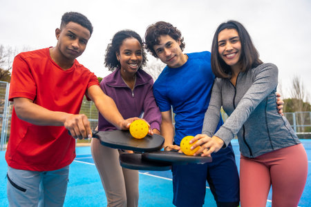 Four happy diverse friends on a bright blue pickleball court, smiling at the camera and holding...
