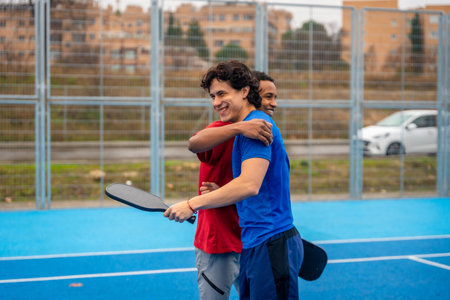 Two diverse male friends smiling and hugging on a blue pickleball court after a game, demonstrating companionship, teamwork, and healthy competition in an urban settingの写真素材