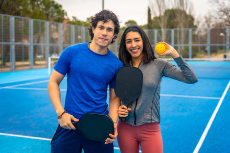 Enthusiastic young couple standing on a blue pickleball court, smiling and holding paddles and a ball, actively participating in a popular growing sport, ready for a healthy workoutの写真素材