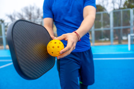 Athlete preparing for a game, holding a perforated yellow pickleball and a black paddle on a blue court, representing a popular racquet sport and active lifestyleの写真素材