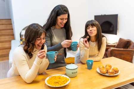 Three young women smiling and laughing, holding cups and spoons, gathering around a wooden kitchen counter with muffins and cereal for a friendly morning social eventの写真素材